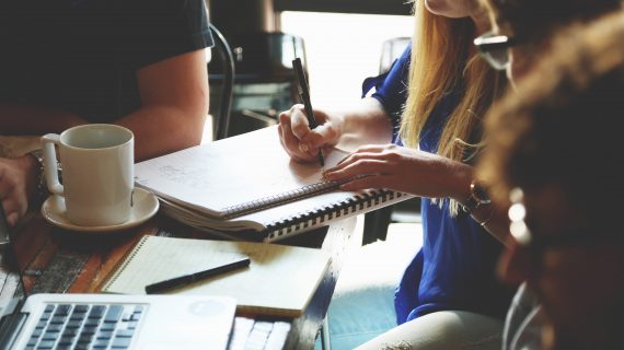 Image of people working at a coffee shot, one woman is writing on a notebook, while another is taking notes