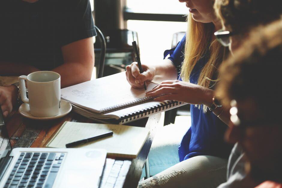 Image of people working at a coffee shot, one woman is writing on a notebook, while another is taking notes